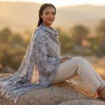 Minimalist antique ivory pashmina shawl with intricate charcoal grey jacquard paisley patterns draped over a model in a sunset desert landscape.