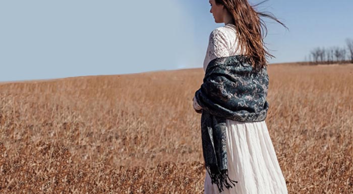 Rear view of a woman standing in a golden wheat field wearing a white lace dress and a dark charcoal grey paisley pashmina shawl with tassel fringe.
